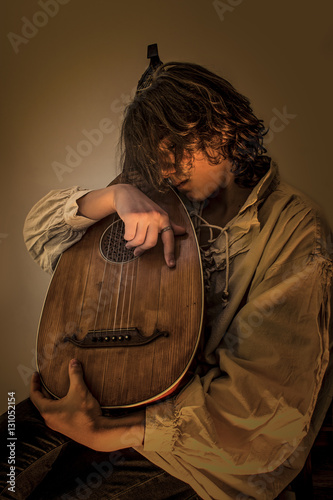 Young man with long blond hair sitting in dark environment and hold in His Hands and hug an Old Oud, Guitar Lute. He is dressed in white rustic baroque shirt with ruffle edging to collars. 