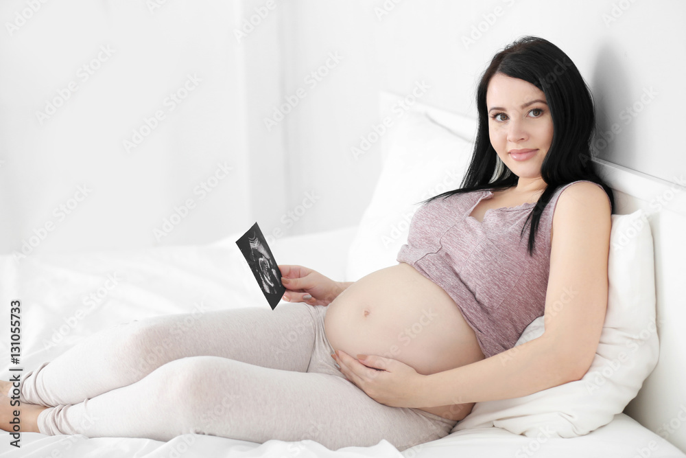 Pregnant woman looking at ultrasound photo and sitting on bed in the room Pregnant woman looking at ultrasound photo and sitting on bed in the room