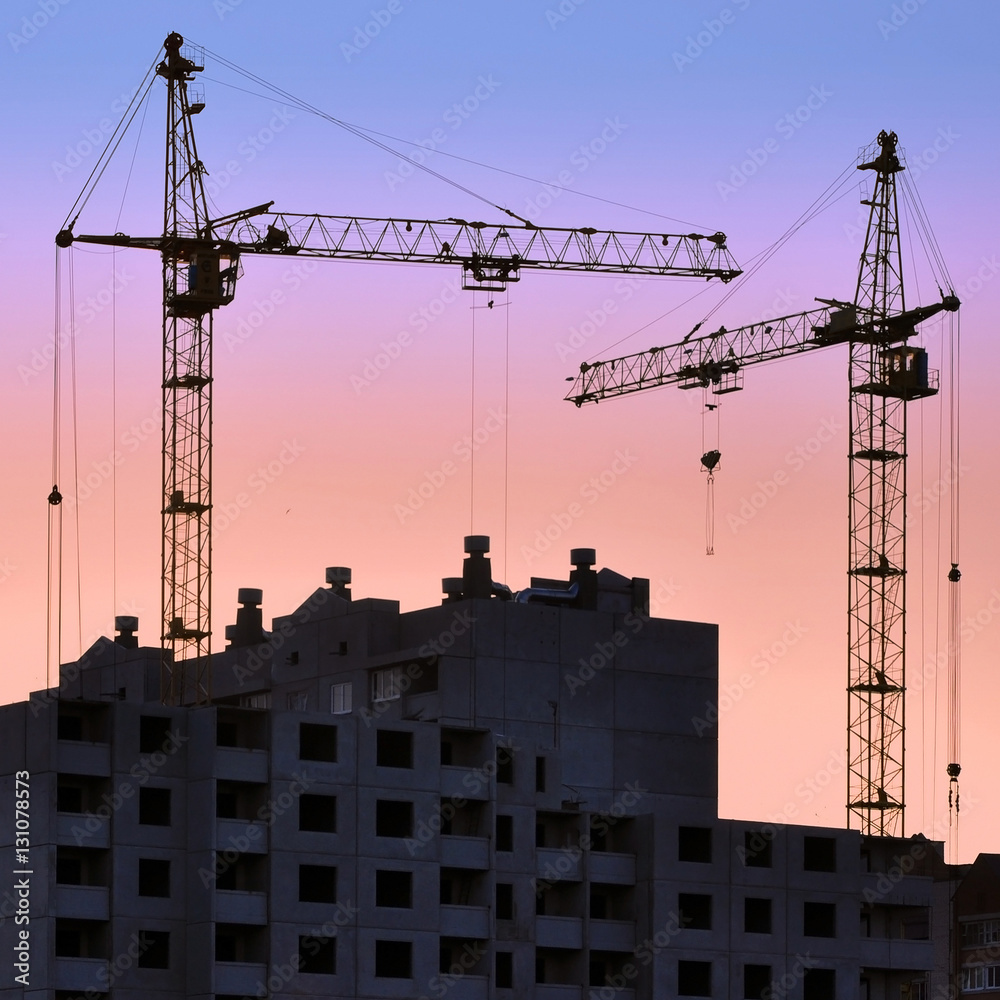 Two construction cranes and building house against the sunset.