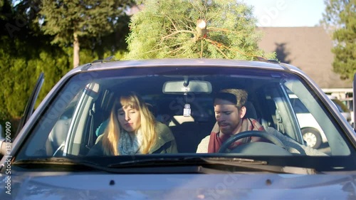 Young man and woman get into their car with baby in the back seat and Christmas tree on top