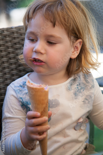 portrait of two years old blonde cute child with white shirt sitting and biting strawberry ice cream cone at urban park  