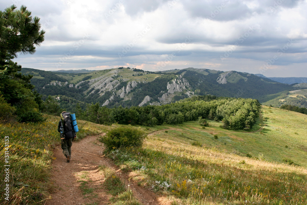 The man with the backpack go on the path in mountains on the summer sunny day. This photo was taken in Crimean Mountains, on South Demerdzhi mountain.