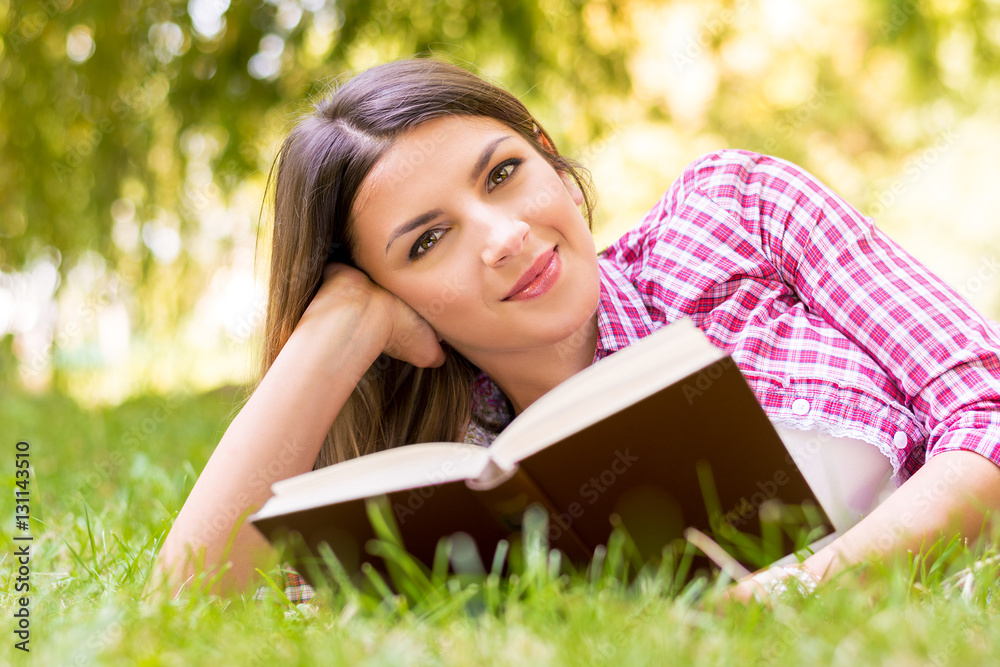Obraz premium Smiling beautiful young woman lying on grass and reading book