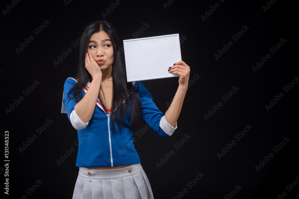 Frightened Asian woman holding exercise book isolated on black. Pretty lady with red lips posing with blank poster in studio. Frightened Asian woman holding exercise book isolated on black. Pretty lady with red lips posing with blank poster in studio.