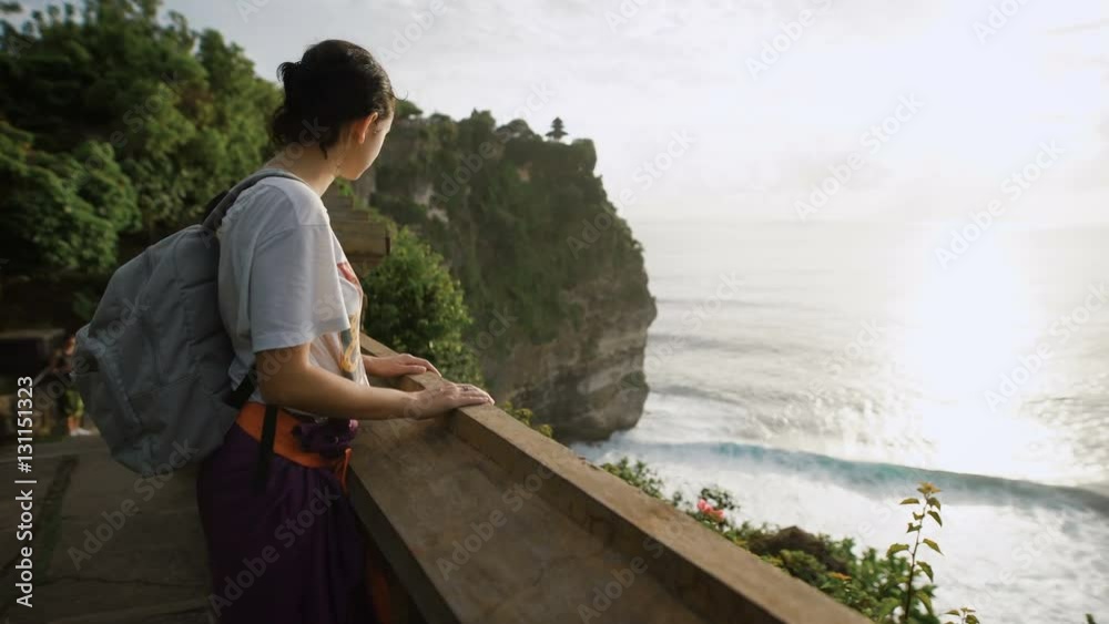 Female tourist standing on the top of mountain with breathtaking view of the ocean and cliff. Girl is looking at the endless water surface of the sea from high point of touristic route in Bali.