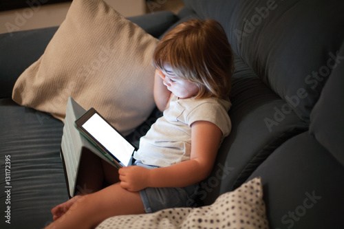 blonde three years old baby shirt and shorts, sitting comfortably in sofa inside home at night reading and watching blank screen digital tablet, face illuminated
