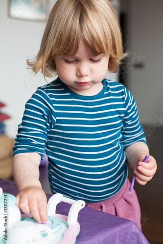 blonde two years old child with striped blue and white sweater inside home playing with plasticine and toys on purple table 