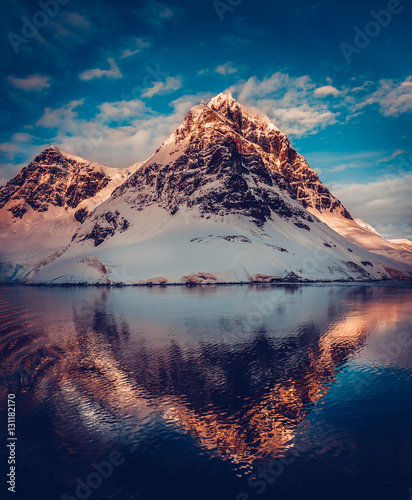 Antarctic landscape with snow covered mountains reflected in ocean water. Sunset warm light on the mountain peak, blue cloudy sky in the background. Exploring beauty world