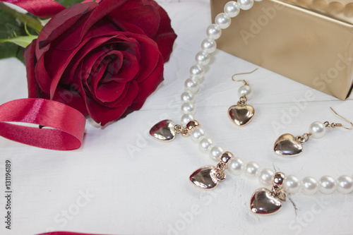 Pearl necklace and earrings with golden gift, red rose and red ribbon on white wood