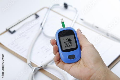 Hand holding a blood glucose meter measuring blood sugar, the background is a stethoscope and chart file