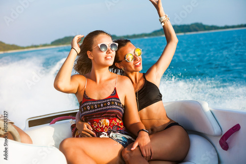 Two pretty amazing hipster girls, blond and brunette ride on a speed boat, wearing sunglasses, happy, tanned, crazy emotions, sexy bikini, fashion accessories fashion, luxury, lifestyle waves splashes photo