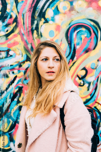 Half length portrait of young beautiful caucasian blonde hair woman leaning on a colorful wall, overlooking smilng - carefree, serene, thoughtless concept