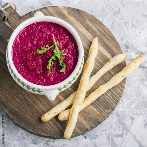 Spring light fresh beet cream soup in portion bowl on a wooden board for feeding with bread sticks. Top view