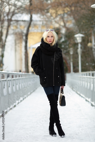 Winter portrait: young blonde woman dressed in a warm woolen jacket blue jeans long boots posing outside in a snowy city park