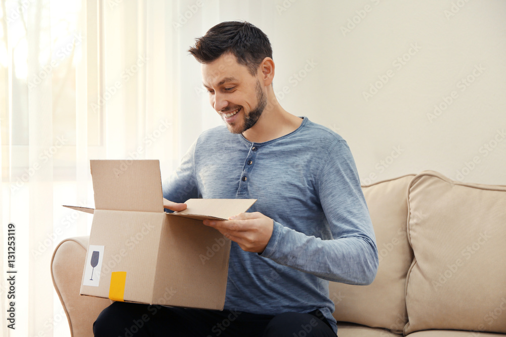 Handsome young man opening box with parcel while sitting on sofa at home Handsome young man opening box with parcel while sitting on sofa at home