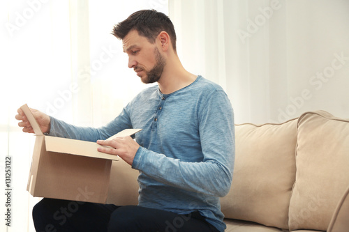 Handsome young man opening box with parcel while sitting on sofa at home