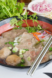 A bowl of traditional vietnamese beef and chicken pho street food, surrounded by fresh herbs. Pho is a vietnamese noodle soup consisting of meat, herbs, chilli, coriander and dumplings.