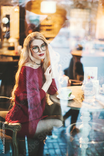 Beautiful young blond woman with glasses sitting in a cafe and drink coffee  it can be seen through the cafe window