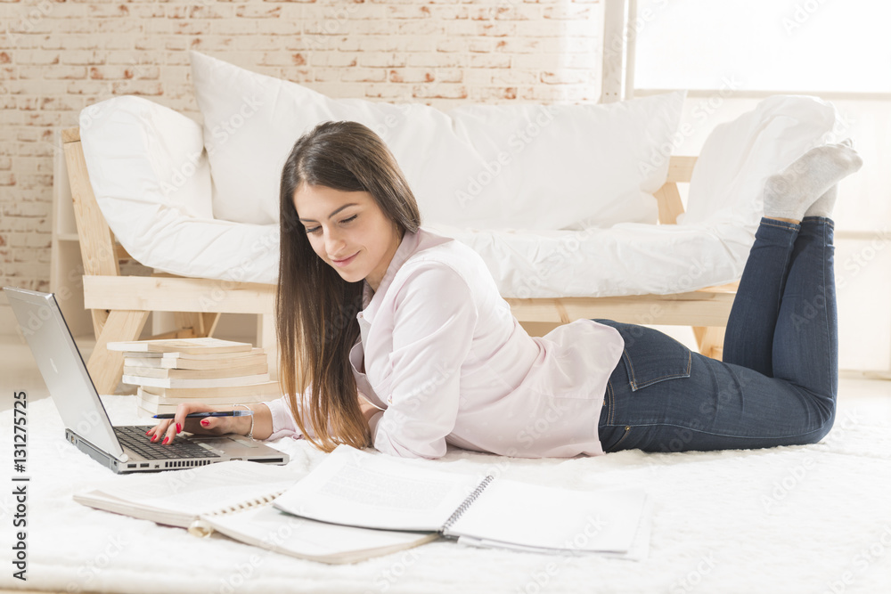Smiling young woman laying down on the floor typing on laptop, dressed in casual clothes. Online study. Work from home