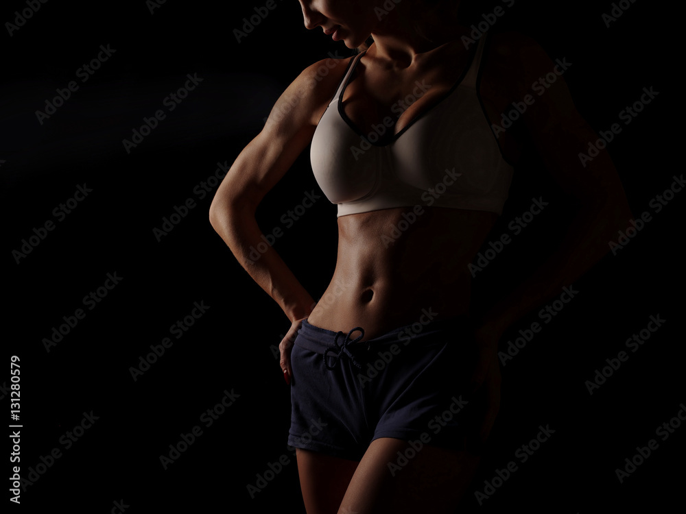 Young athletic and sexy woman in a white sports shirt and blue shorts in the studio on a black background shows strong biceps and a flat belly with the perfect abdominal muscles, dark black background Young athletic and sexy woman in a white sports shirt and blue shorts in the studio on a black background shows strong biceps and a flat belly with the perfect abdominal muscles, dark black background