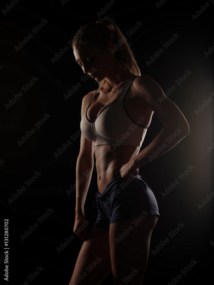 Beautiful athletic young woman with slim muscular body (perfect abdominal muscles, strong biceps, sexy buttocks) wearing the sportswear is posing on the dark black studio background, diet and fitness