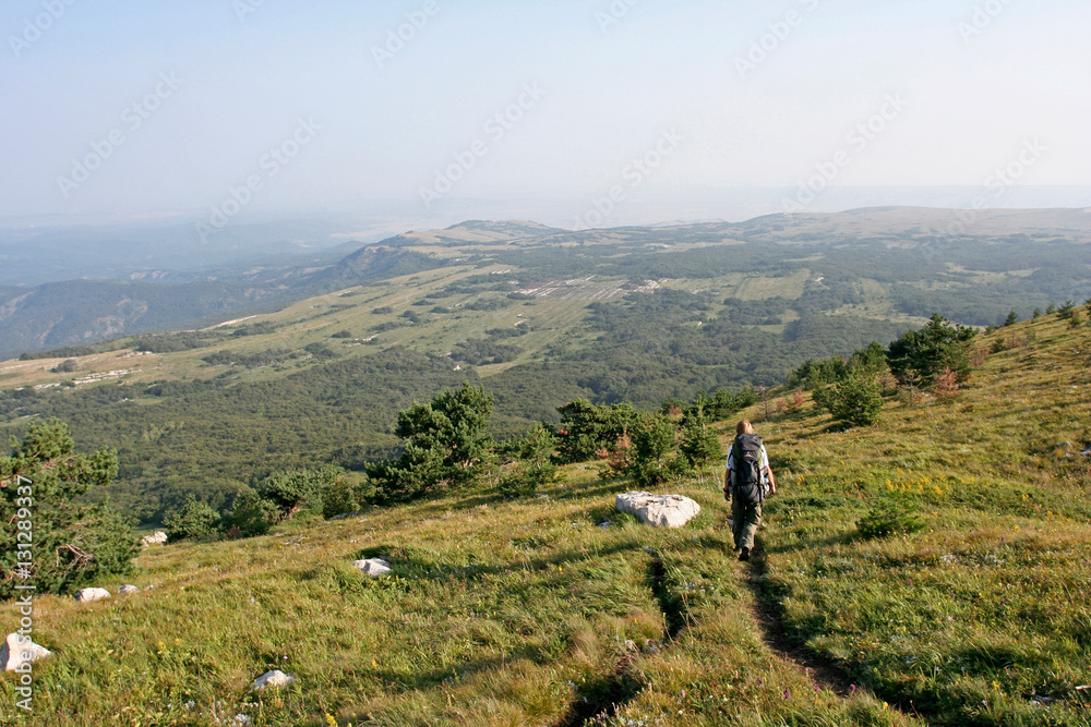 The man with the backpack go on the path in mountains on the summer sunny day. This photo was taken in mountainous massif Chatyr-Dag, Crimea.