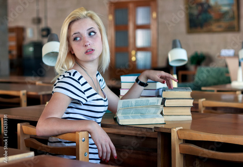 Tired girl coquettishly turns around to look back. She sits at a table on which there are a pile of books and reading lamps photo