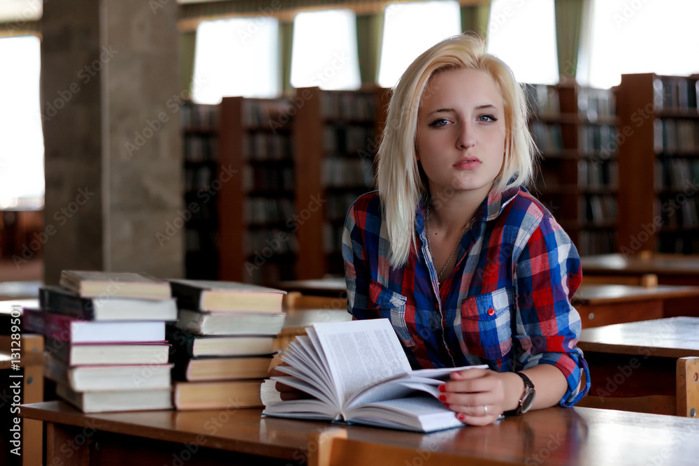 Young blonde woman sitting at the table. In the background are the rows of bookshelves. She holds in her hands an open book. Her haughty gaze directed at the camera Young blonde woman sitting at the table. In the background are the rows of bookshelves. She holds in her hands an open book. Her haughty gaze directed at the camera