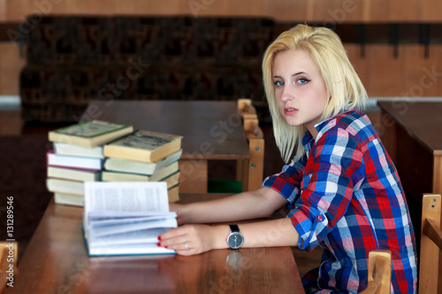 Young blonde girl in a plaid shirt sitting at desk, holding an open book in his hands. Side view.