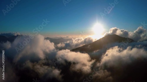 Aerial bird view flight backward over clouds flying away from sun showing blue sky background and mountain landscape below beautiful scene looks like heaven's gate person of view first person view 4k photo