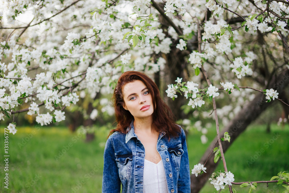 Young pretty serious caucasian woman in jeans jacket near blooming spring tree and green grass. Youth, freshness, beauty, happiness concept. Young pretty serious caucasian woman in jeans jacket near blooming spring tree and green grass. Youth, freshness, beauty, happiness concept.