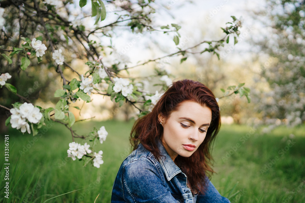 Young pretty serious caucasian woman in jeans jacket near blooming spring tree and green grass. Youth, freshness, beauty, happiness concept. Young pretty serious caucasian woman in jeans jacket near blooming spring tree and green grass. Youth, freshness, beauty, happiness concept.