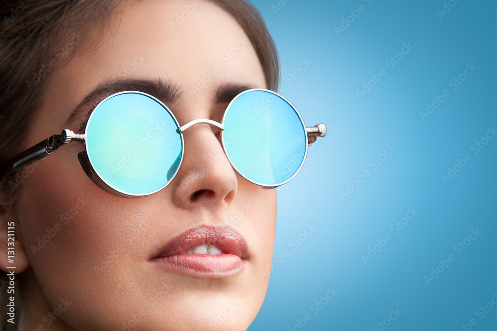 Close-up face portrait of young beautiful woman with perfect skin in round sunglasses looking up on blue background. Beauty face make-up. Close-up face portrait of young beautiful woman with perfect skin in round sunglasses looking up on blue background. Beauty face make-up.