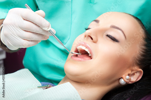 Close-up of young beautiful woman patient with perfect straight white teeth sitting in dentist chair while doctor examines her teeth with the help of dental mirror. Healthcare, medicine.