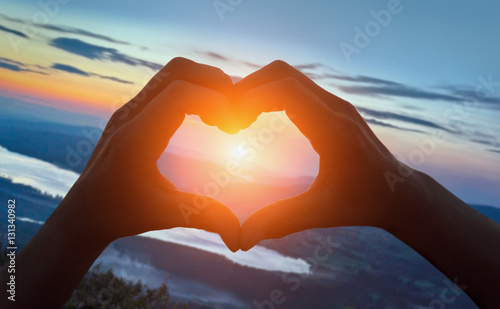 Hands making heart shape on the beach against sunrise over mount