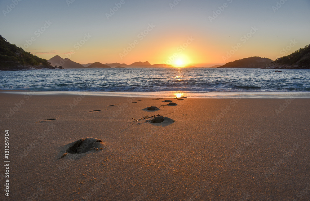 Beautiful sunrise with the sun rising from the Atlantic Ocean, solar path on the water and footprints at the deserted Praia Vermelha Beach, Rio de Janeiro, Brazil