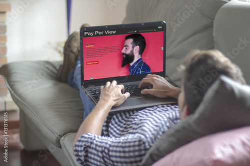 Young man lying on the sofa using laptop with pay per view website on the screen. View from behind. All screen graphics are made up. photo