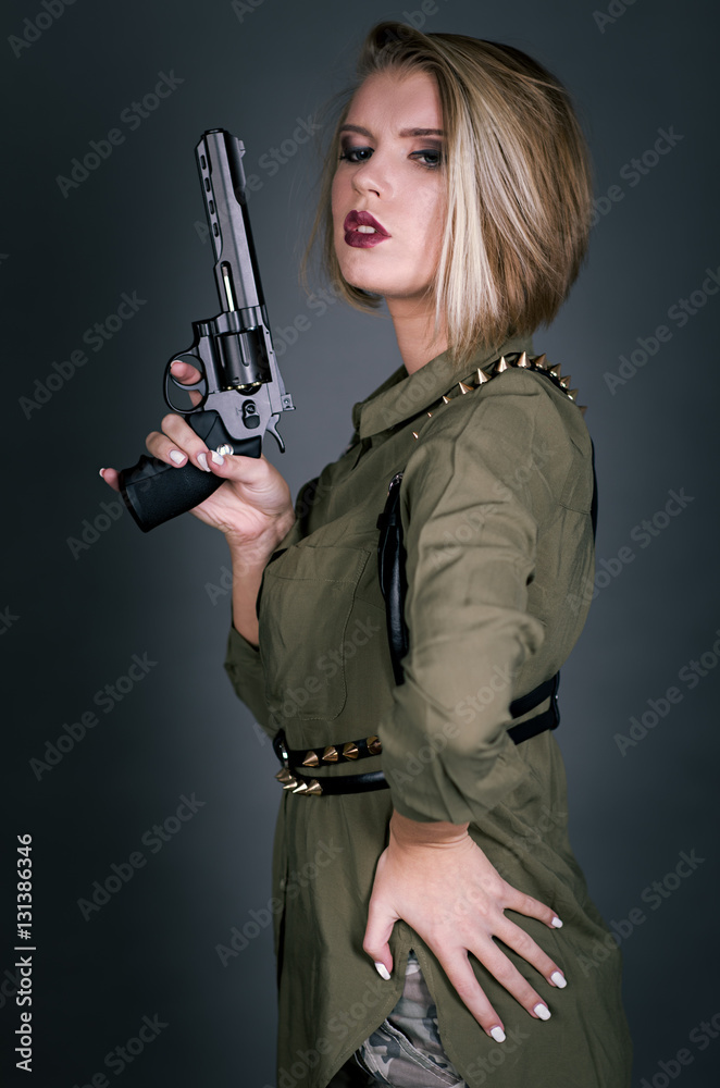 Half-turn portrait of young blonde woman dressed in military style shirt and belt holding handgun close to face Half-turn portrait of young blonde woman dressed in military style shirt and belt holding handgun close to face