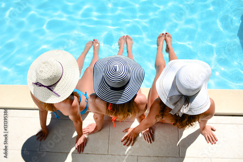 Photo three beautiful young woman with sun hat sitting by the poolside of a resort swi