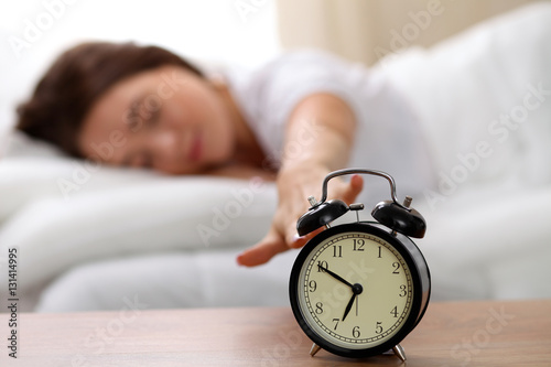 Sleepy young woman stretching hand to ringing alarm willing turn it off. Early wake up, not getting enough sleep, getting work concept.
