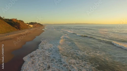 Aerial flying shot down a beach in California over the sand and water. Beach houses in the distance. Sunset time with clear blue skies and golden light. 