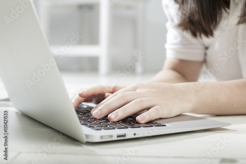 Girl in light colored clothes typing using hands on gray laptop keyboard, lying on a white floor