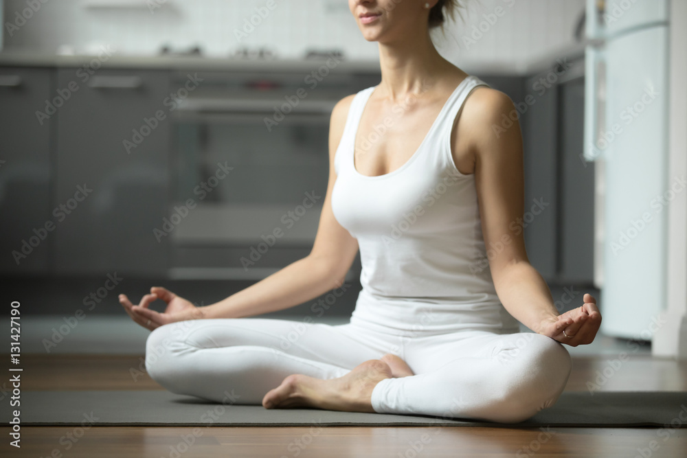 Sporty attractive woman practicing yoga, sitting in Half Lotus exercise, Siddhasana pose, working out, wearing white sportswear, indoor, home interior background. Midsection close up Sporty attractive woman practicing yoga, sitting in Half Lotus exercise, Siddhasana pose, working out, wearing white sportswear, indoor, home interior background. Midsection close up