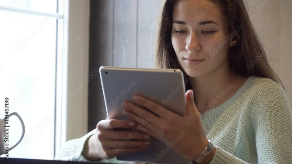 Young brunette girl sits on the table near window and work on tablet pc Young brunette girl sits on the table near window and work on tablet pc
