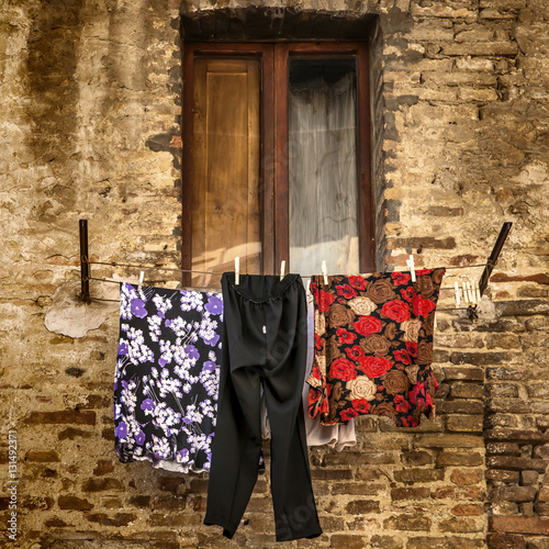 Close crop of colourful washing hanging on a line in front of a half shuttered window set in an old brick wall of a Tuscan village, square format  © William