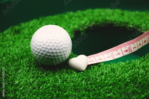 golf ball and white heart shape on green grass at hole with measuring tape.