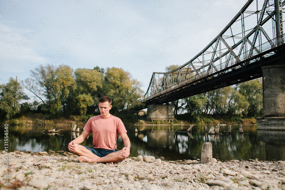 man practices yoga on the river bank near the old bridge man practices yoga on the river bank near the old bridge