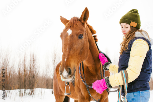 Woman in winter clothes and the horse to stand together against the white snow. love and care for horses. girl puts a bridle on a horse © evgeniykleymenov
