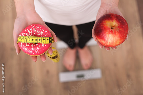 Close up view of woman making choice between apple and donut with blurred scales on background. Dieting concept