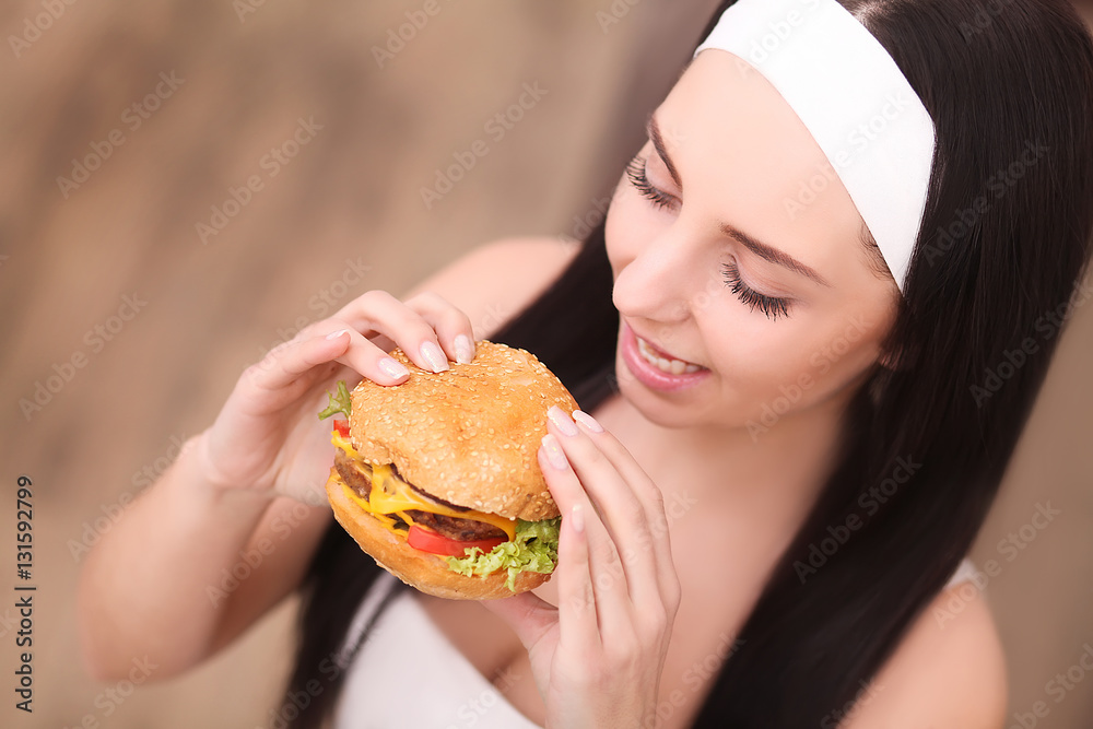 Unhealthy eating. Junk food concept. Portrait of fashionable young woman holding burger and posing over wood background. Close up. Copy-space. Perfect hair, skin, make-up and manicure. Studio shot
